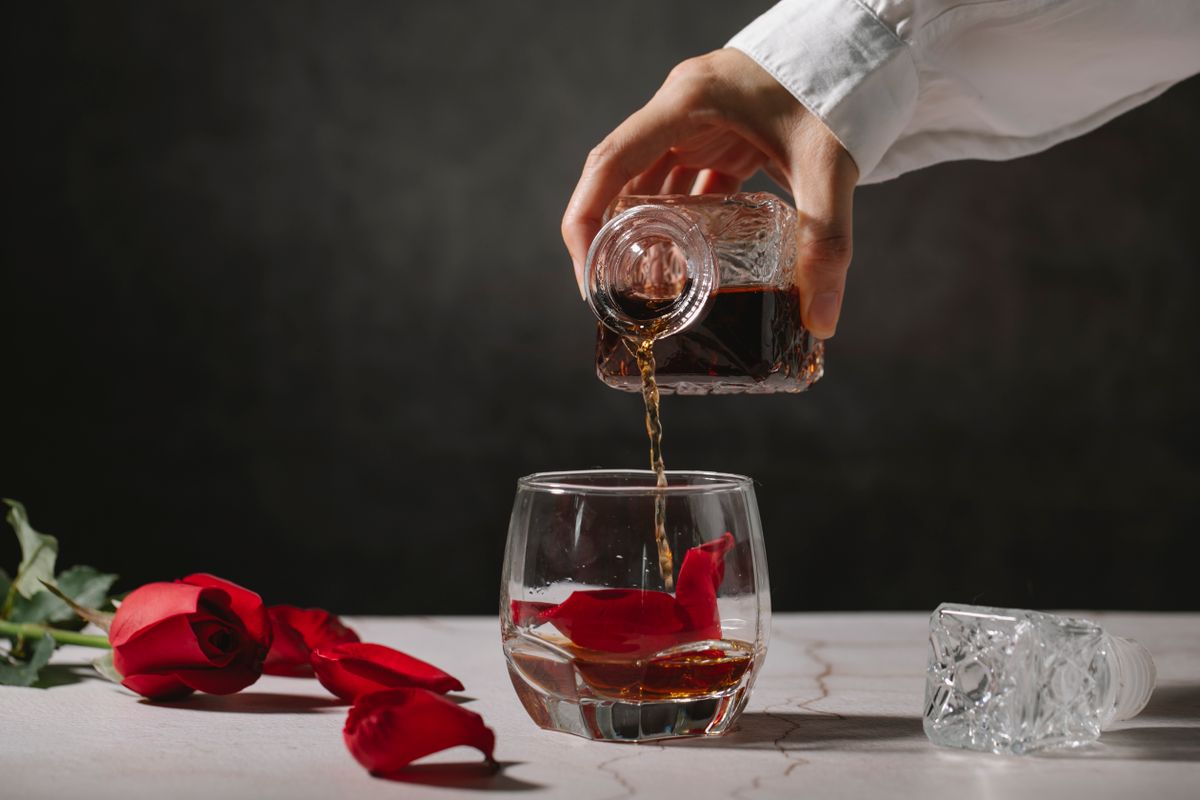 Crop unrecognizable person in white shirt pouring whiskey from crystal jar into transparent glass with petals placed on table near rose flower