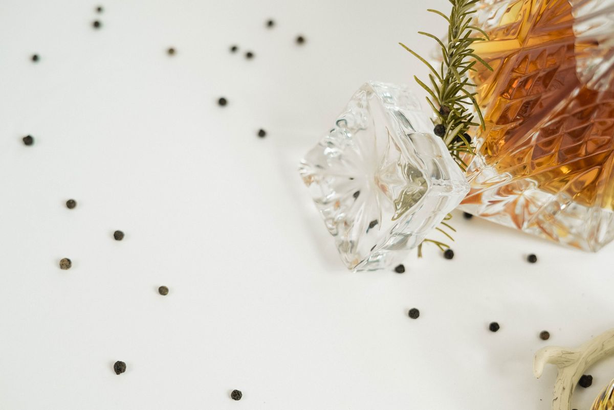From above of glass carafe filled with Scotch whiskey placed on white background with rosemary twig and scattered back peppercorns