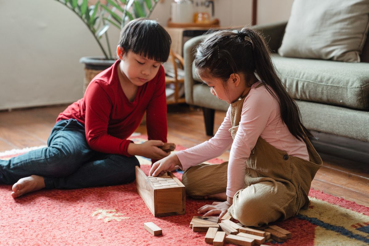 Two children play with wooden blocks on a rug in a cozy living room setting.