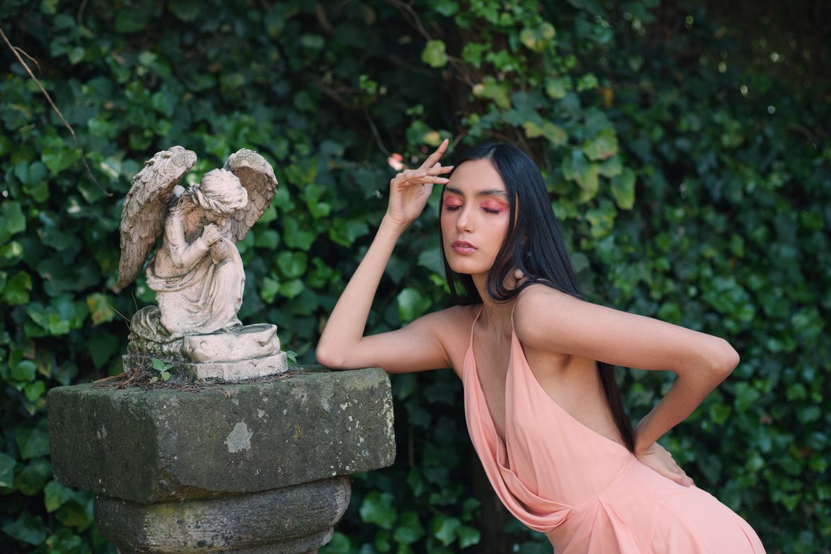Woman in pink dress posing elegantly beside a garden angel statue.
