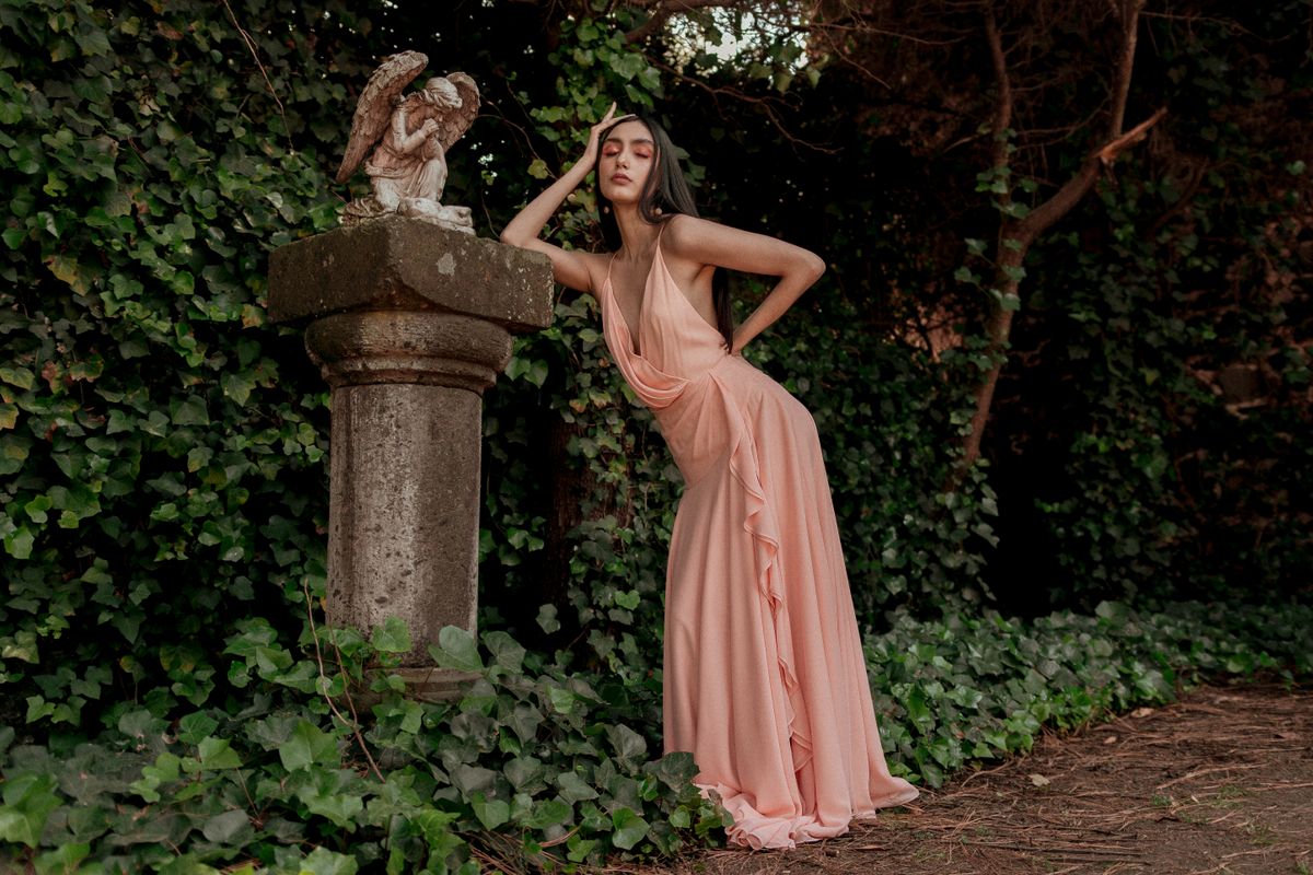 A woman in a peach dress poses by a stone with an angel statue in a lush garden setting.