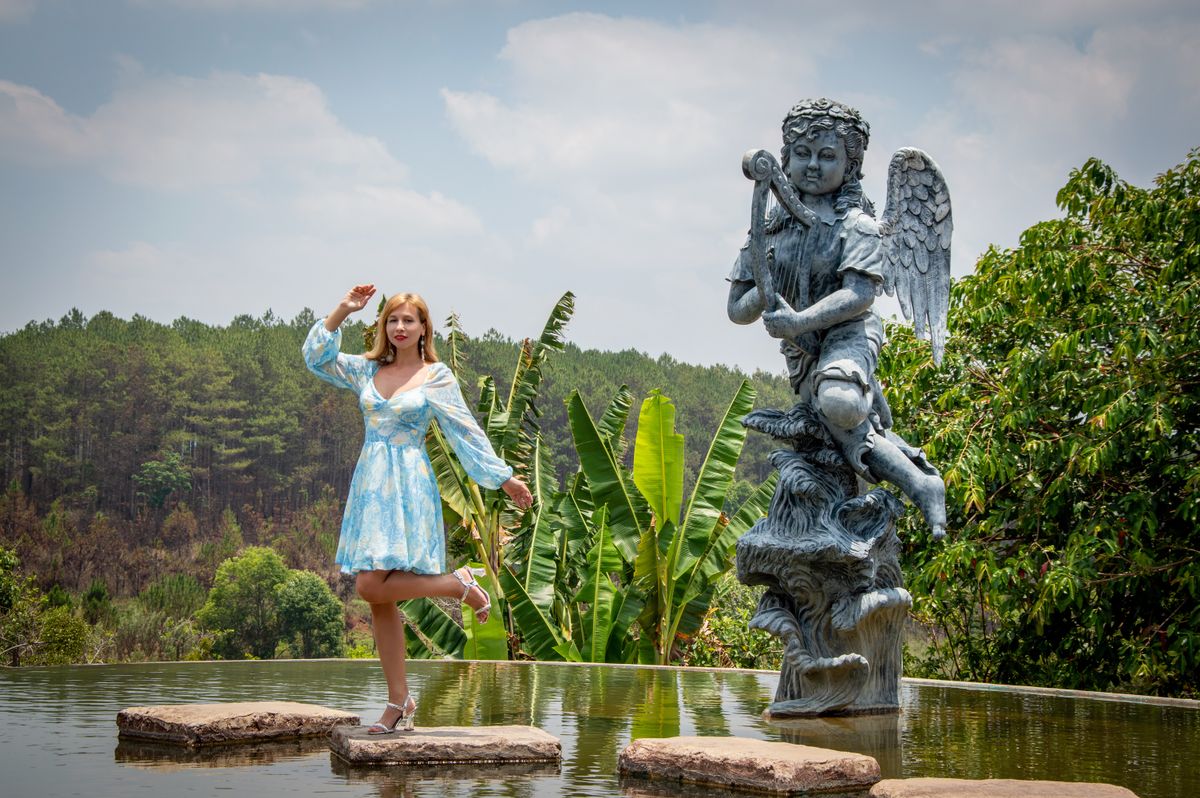 Woman in blue dress posing by angel statue in a lush garden setting.