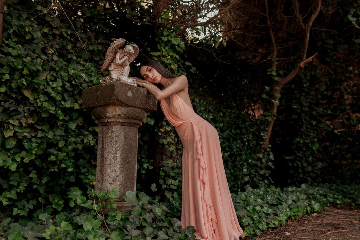 A woman in a peach dress leans on an angel statue in a lush outdoor setting.
