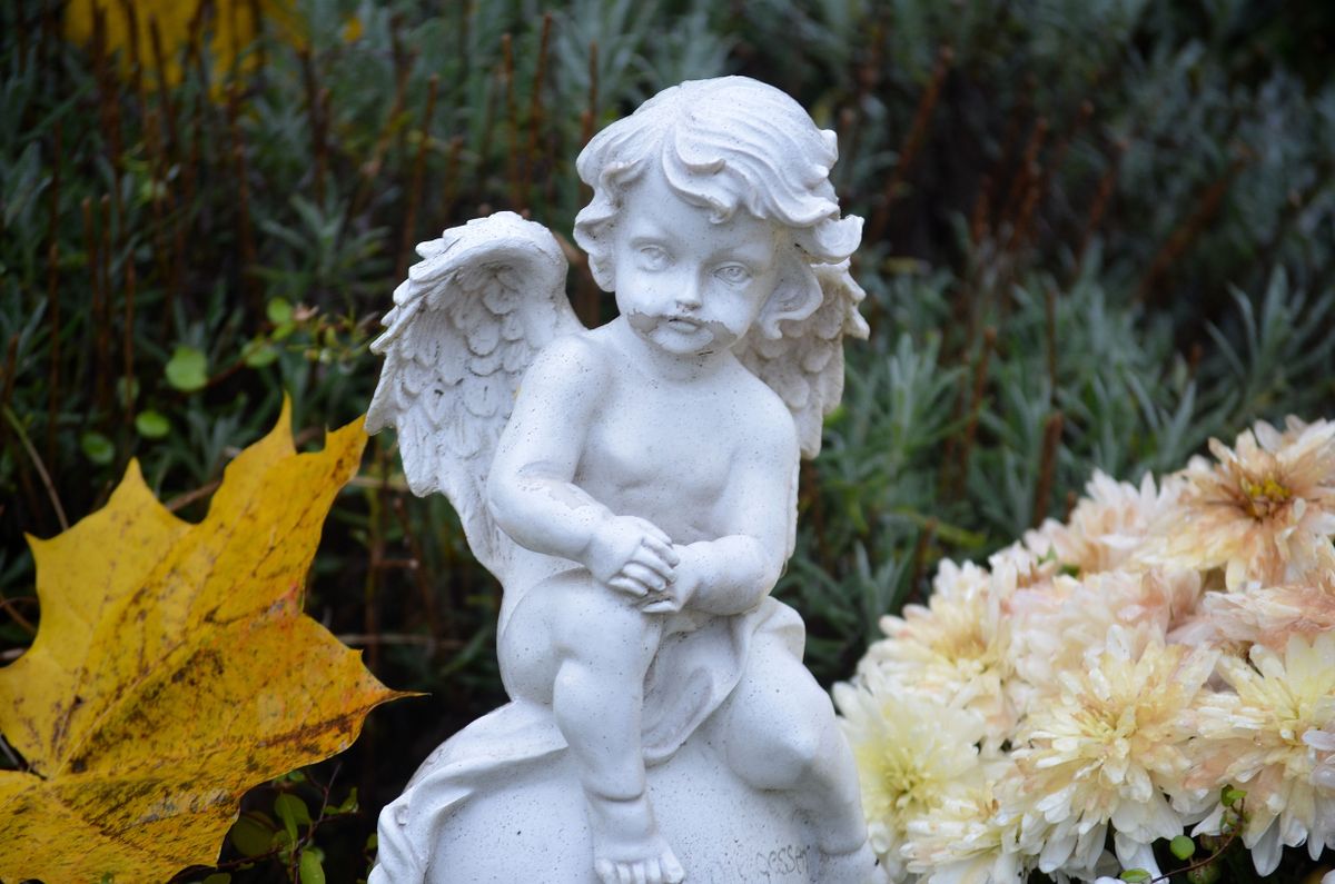 A white stone angel statue amidst autumn leaves and flowers in a garden.