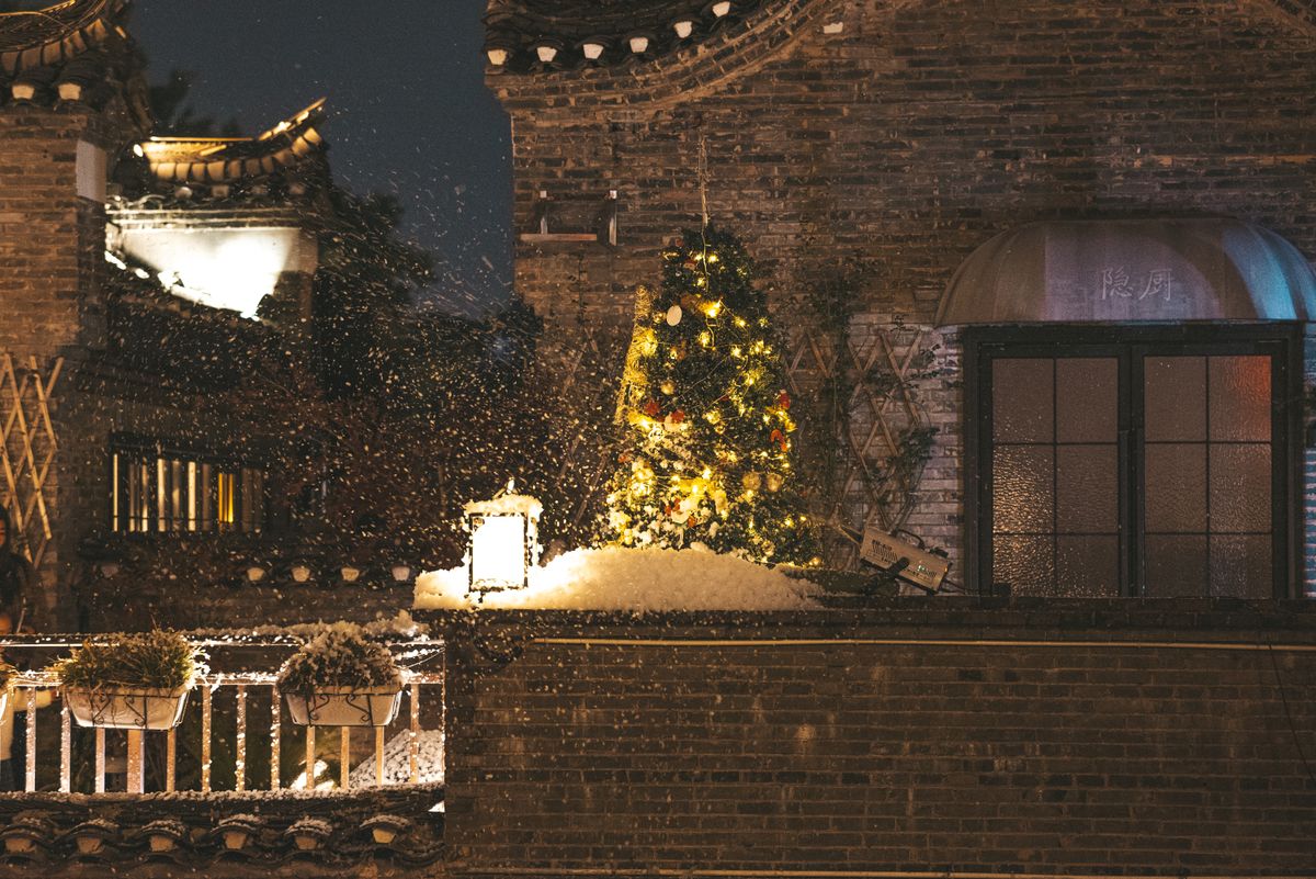 Festive Christmas tree with lights on a snowy rooftop during nighttime snowfall.