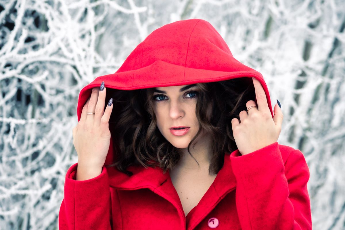 A stylish woman wearing a red hooded coat poses elegantly in a snowy winter setting.