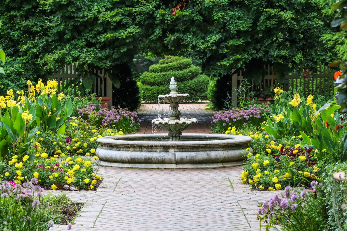 Beautiful, serene garden scene with vibrant flowers and a central stone fountain surrounded by lush greenery.