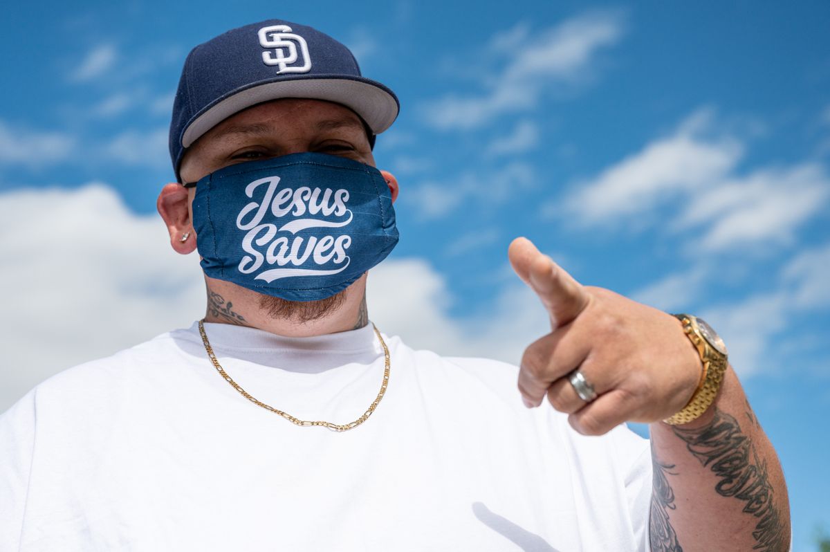 Ethnic man with tattoos in white t shirt and cap and protective mask with Jesus Saves inscription pointing finger at camera