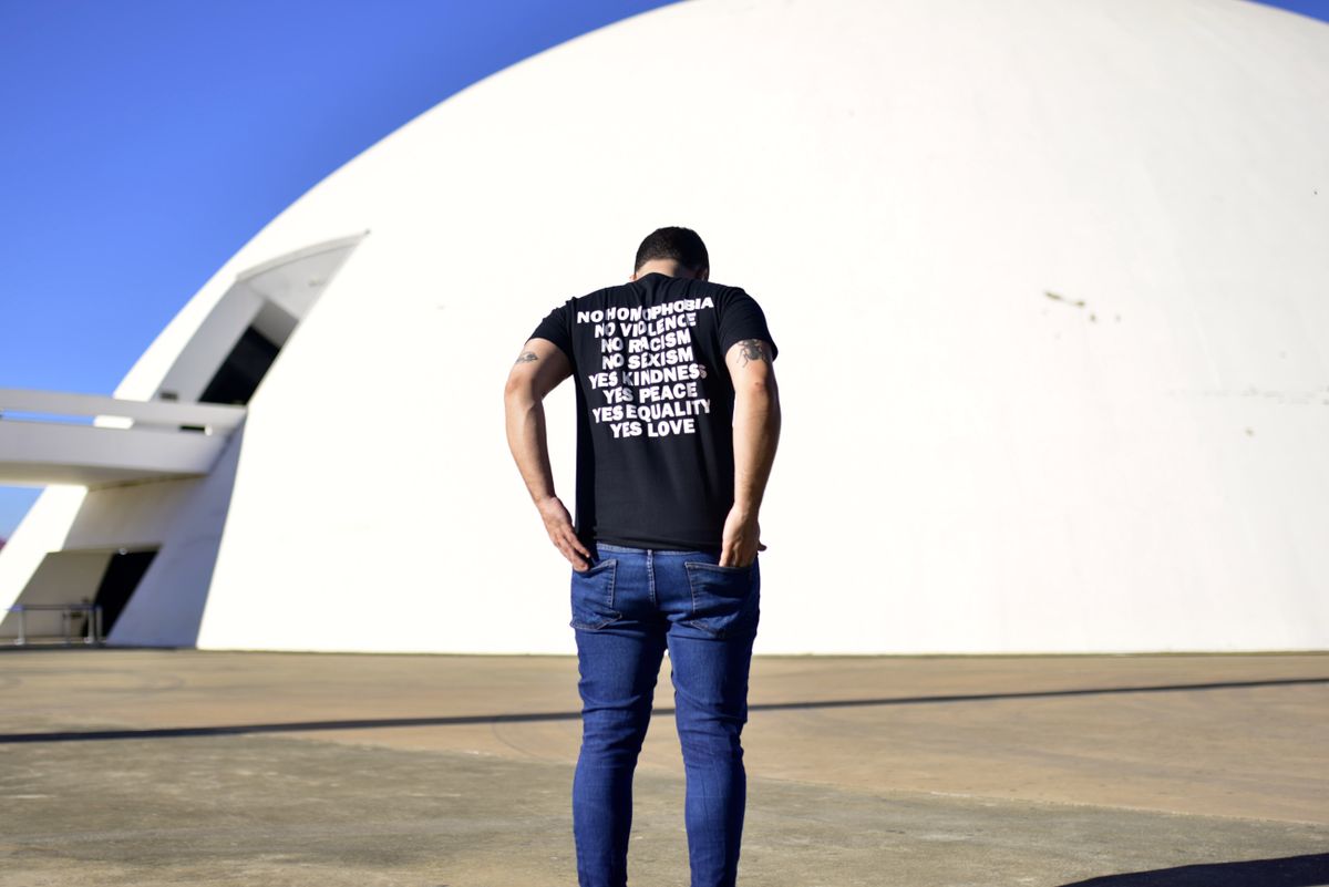 Man stands back to camera wearing a T-shirt with positive messages in front of iconic Brasília architecture.