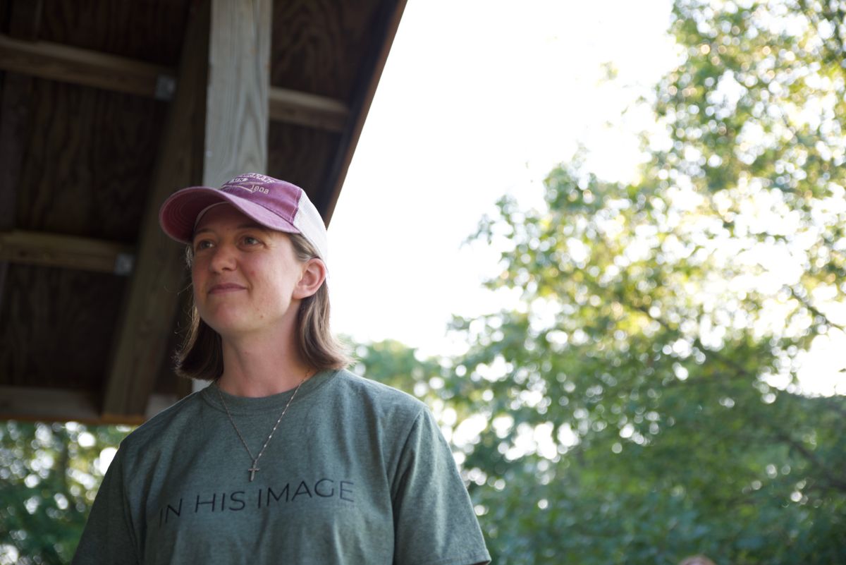 A woman wearing a cap and t-shirt stands outside smiling under a wooden structure.