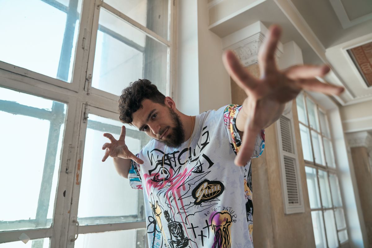 A bearded man poses with expressive hand gestures in a graffiti-style shirt.