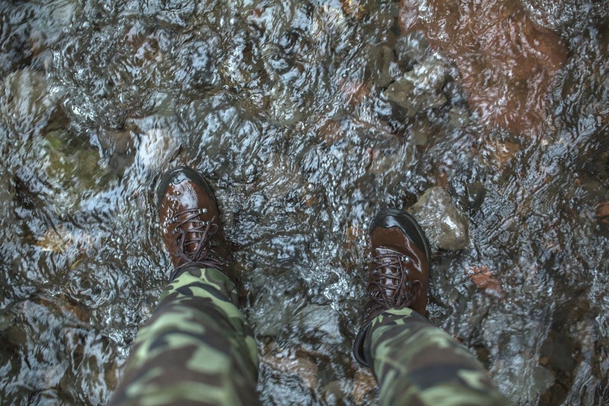 A person standing in a river wearing camouflage pants and brown hiking boots.