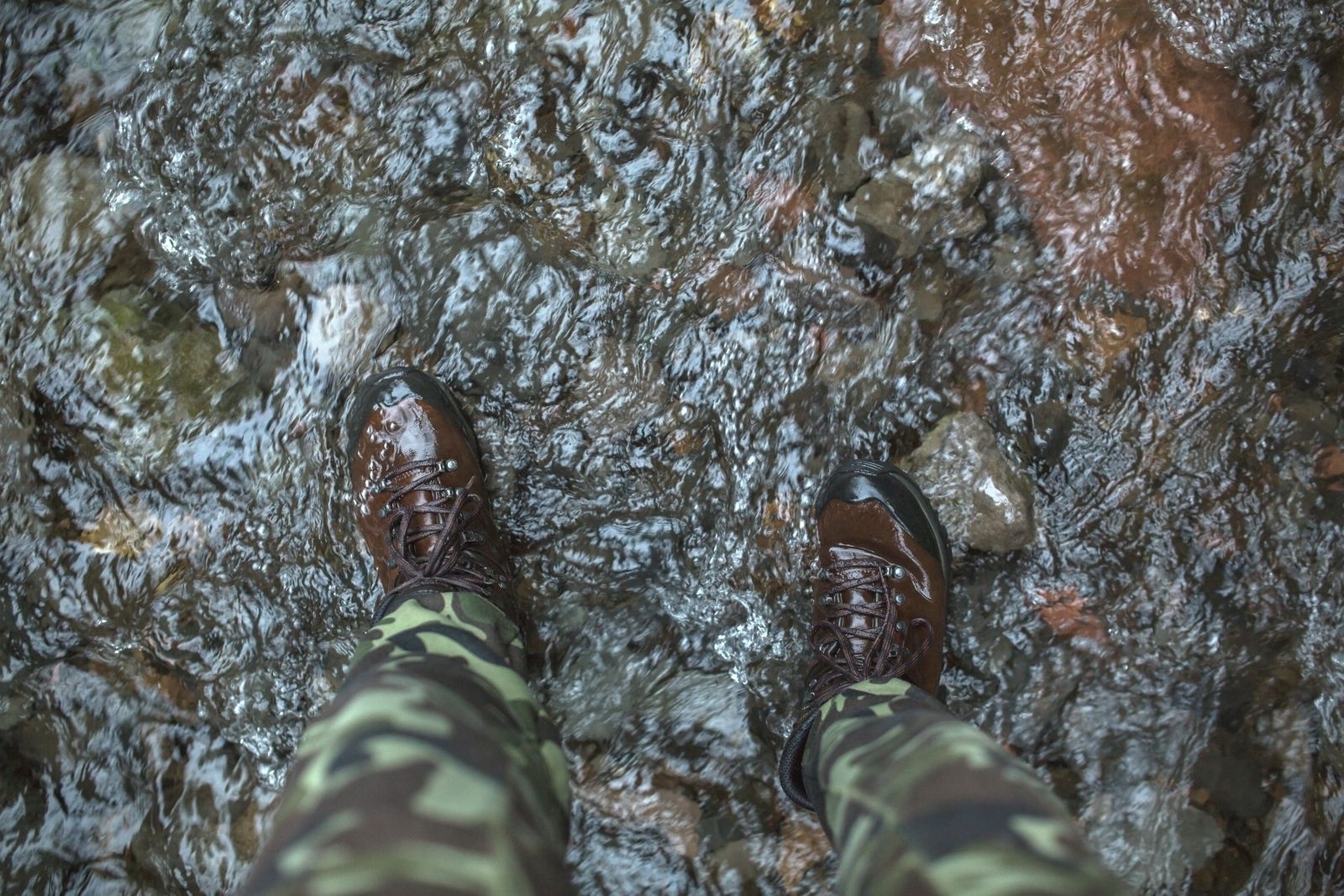 A person standing in a river wearing camouflage pants and brown hiking boots.