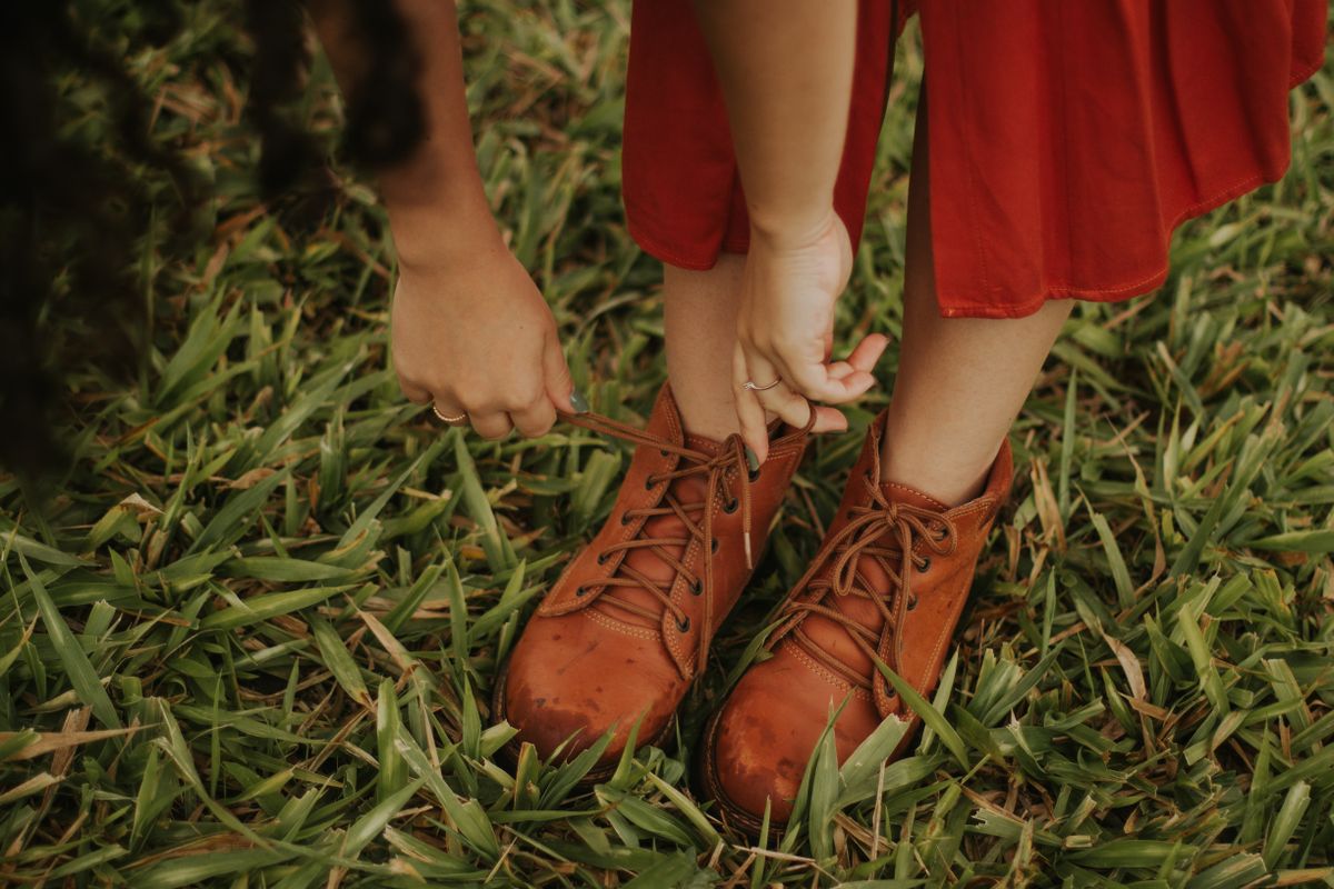A woman tying her brown leather boots on grassy terrain wearing a red dress.