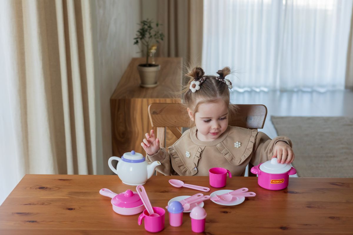 Adorable girl having fun with a pink toy tea set at a wooden table indoors.