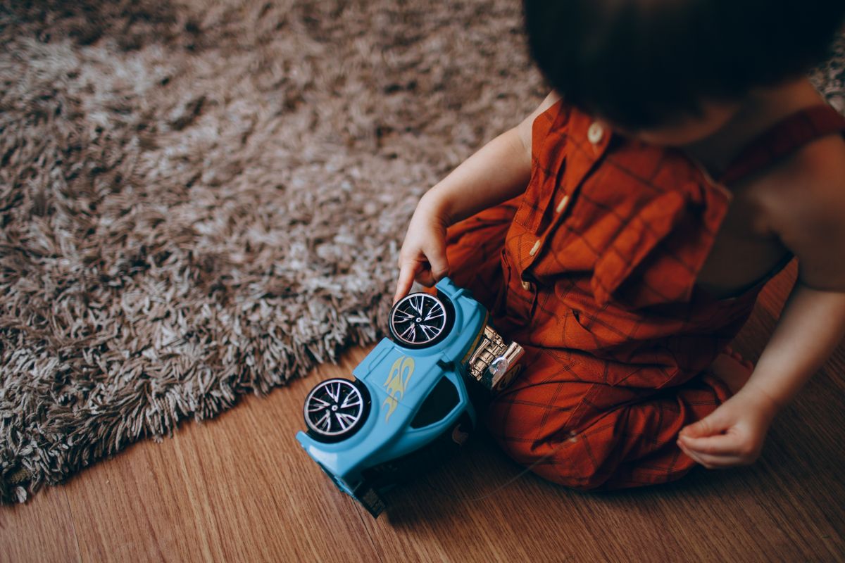 A young child in red overalls playing with a blue toy car on a carpeted wooden floor.