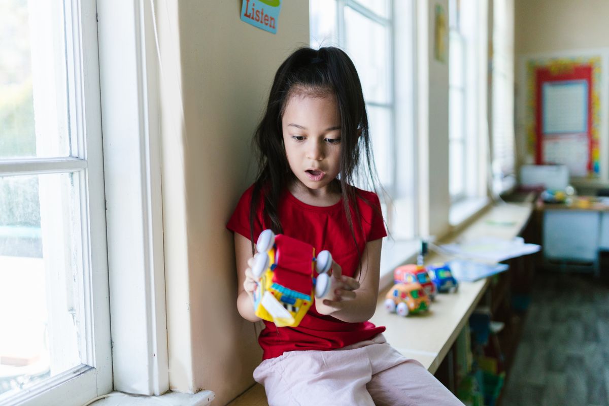 Young girl in red shirt playing with a colorful toy car by a window in a classroom.