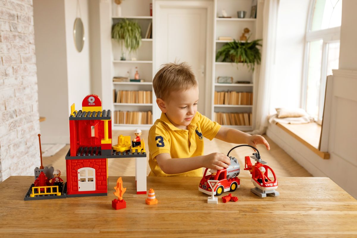 A young boy in a yellow shirt playing with toy building blocks and vehicles on a wooden table.