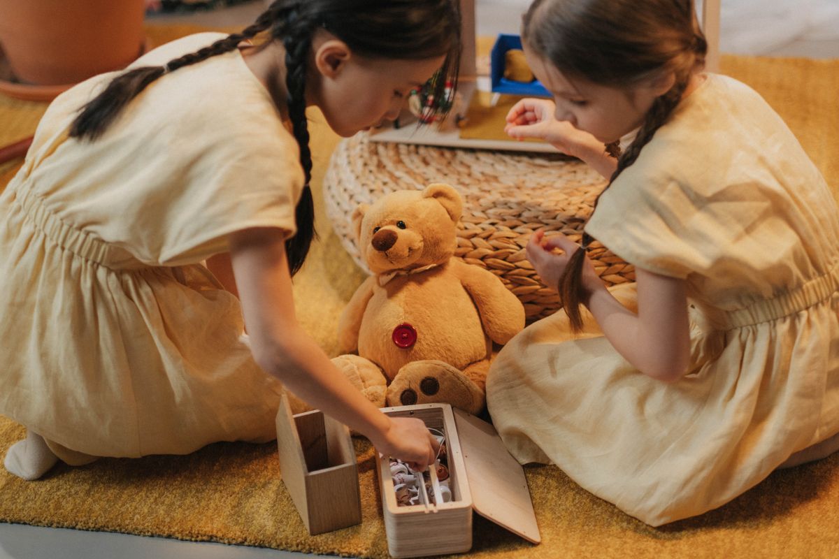 Two girls playing with toys on a woven carpet indoors, featuring a teddy bear and creative imagination.