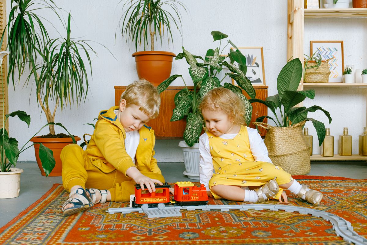 Cute siblings wearing yellow and white clothes sitting on colorful old fashioned carpet and having fun together while playing with plastic railway in living room