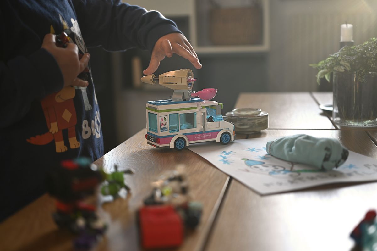 A child plays with a toy van on a well-lit table indoors, surrounded by colored drawings and action figures.
