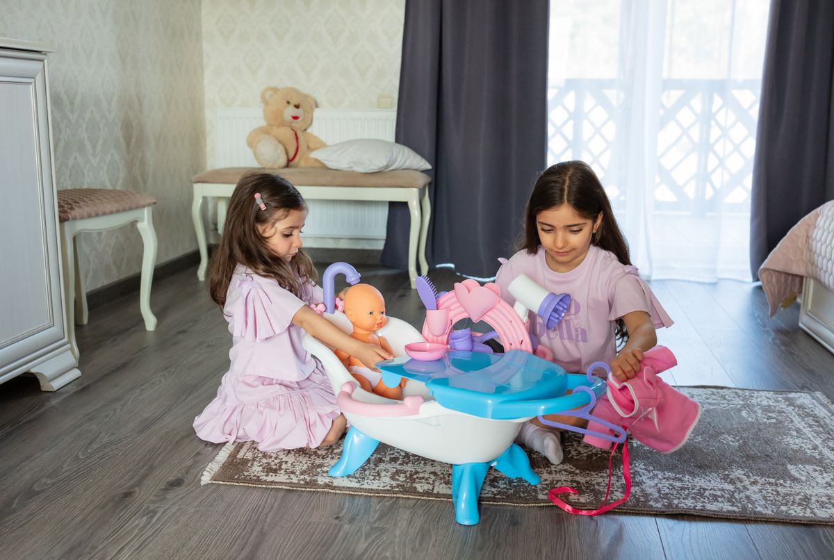 Two young girls engaging in imaginative play with a toy baby and doll accessories indoors.