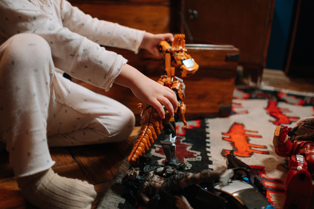 A child in pajamas plays with a toy dinosaur on a colorful rug indoors.