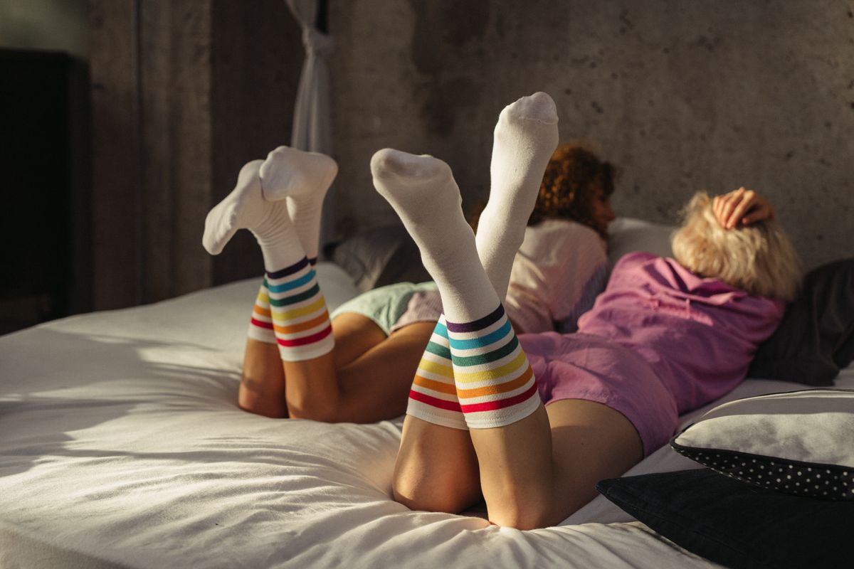 Two friends lounging on a bed, wearing colorful rainbow striped socks, creating a cozy indoor vibe.