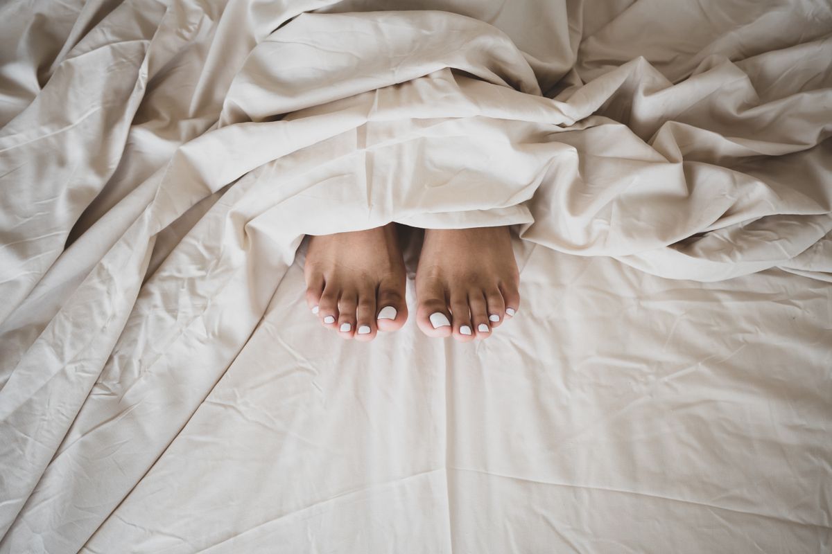 Pair of feet with white nail polish resting on a messy bed, offering a sense of relaxation and comfort.