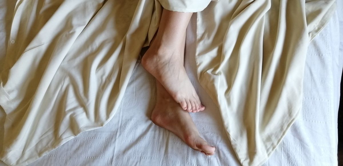 A pair of feet peeks out from under cream-colored linen sheets in a cozy and relaxed setting.