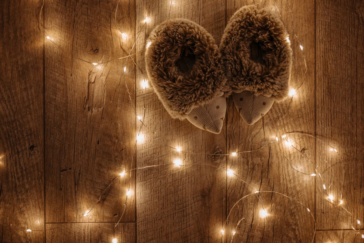 Warm fuzzy slippers surrounded by glowing string lights on a wooden floor, creating a cozy ambiance.