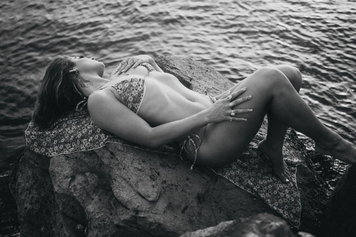 A woman in a bikini relaxes on rocks by the water, captured in black and white.