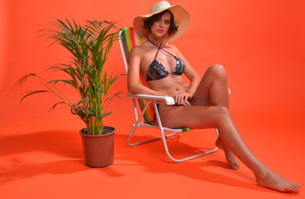 Stylish woman in bikini and sun hat posing on deckchair with potted plant in bright studio.