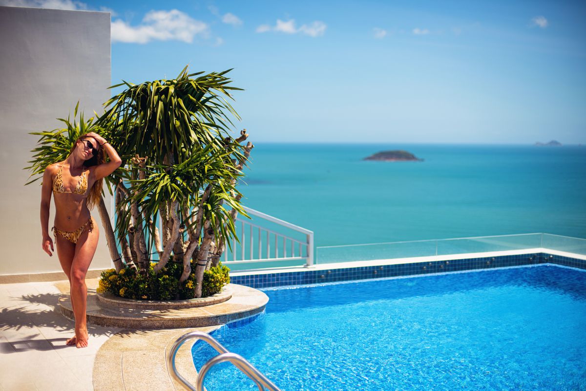 A woman in a leopard bikini stands by a rooftop pool overlooking the ocean.