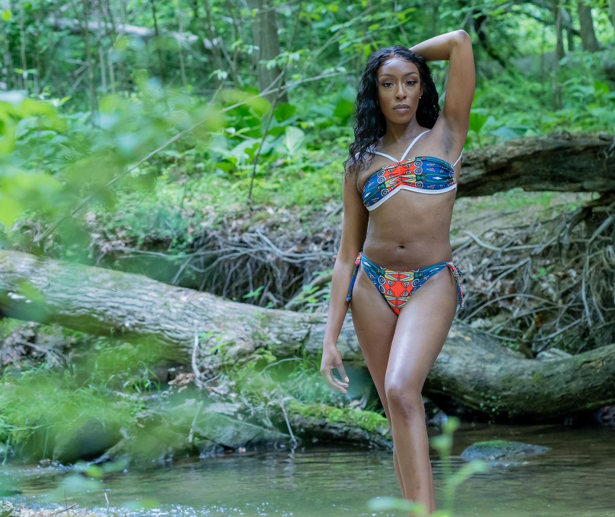 A stunning African American woman poses confidently in a vibrant bikini by a serene riverbank amidst lush greenery.