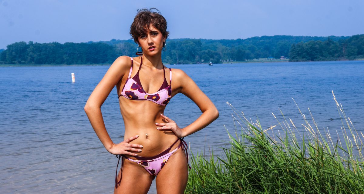 A woman in a floral bikini poses confidently by a serene lakeside setting.