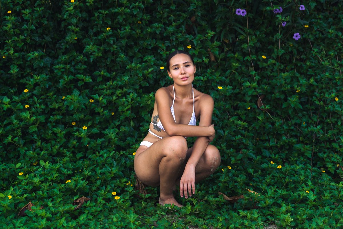 Portrait of a woman in a bikini crouching amidst lush green foliage with yellow flowers.