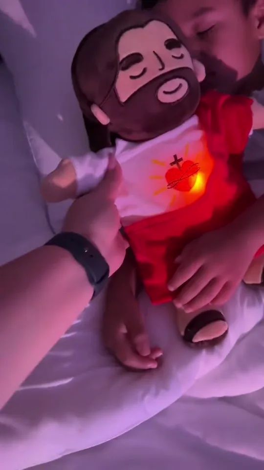 A child asleep in a dark bedroom holding the plush, its Sacred Heart glowing warm