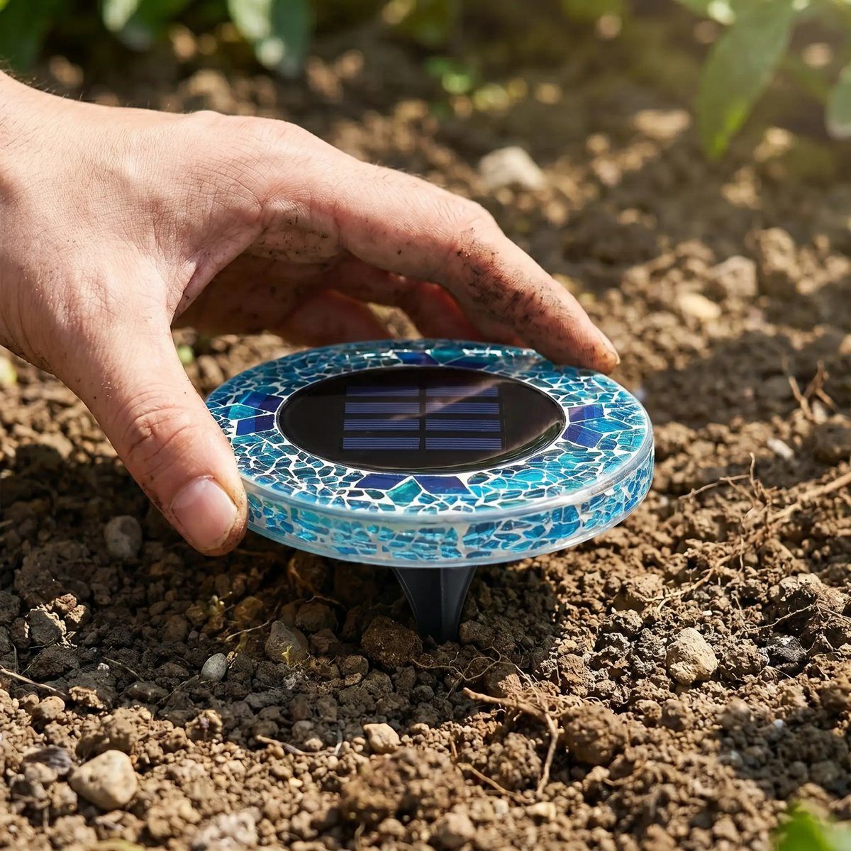 Hand placing a blue mosaic solar disk light into garden soil