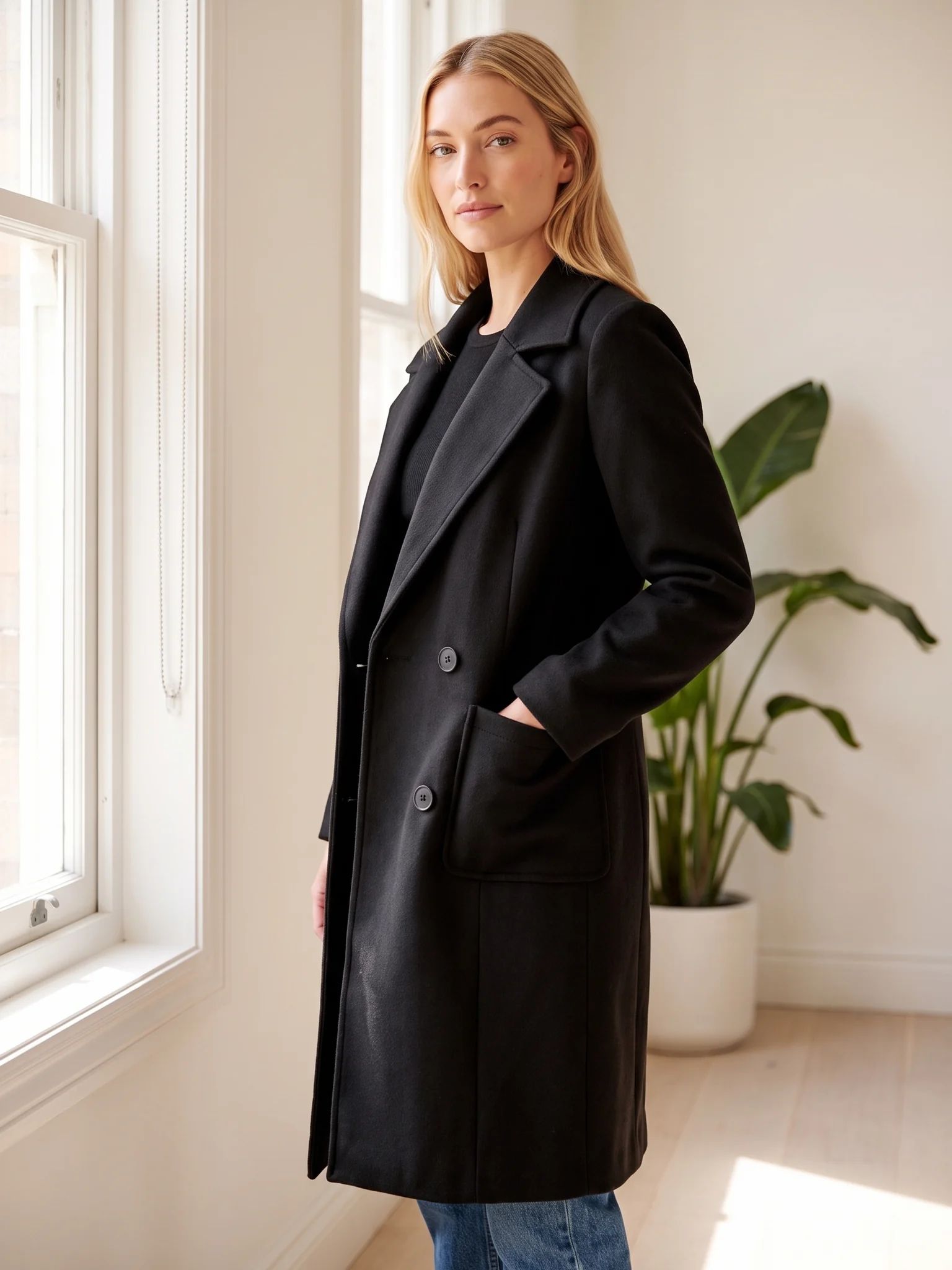 A woman in the Black wool-blend coat standing by a window in a bright apartment